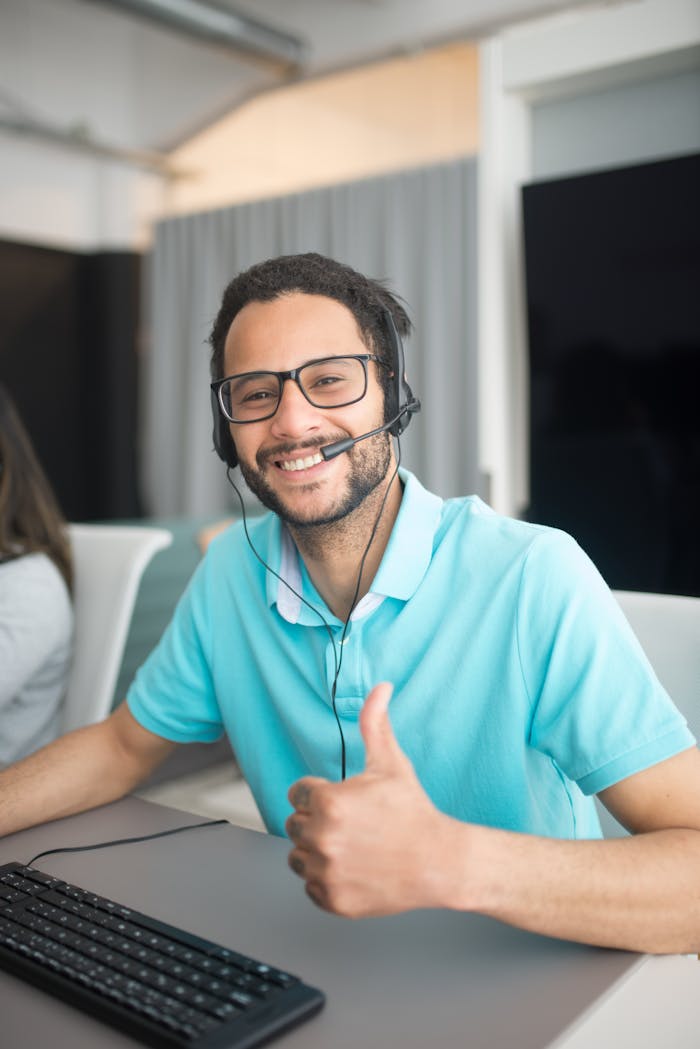 Friendly male call center agent wearing headset, giving thumbs up and smiling in office setting.